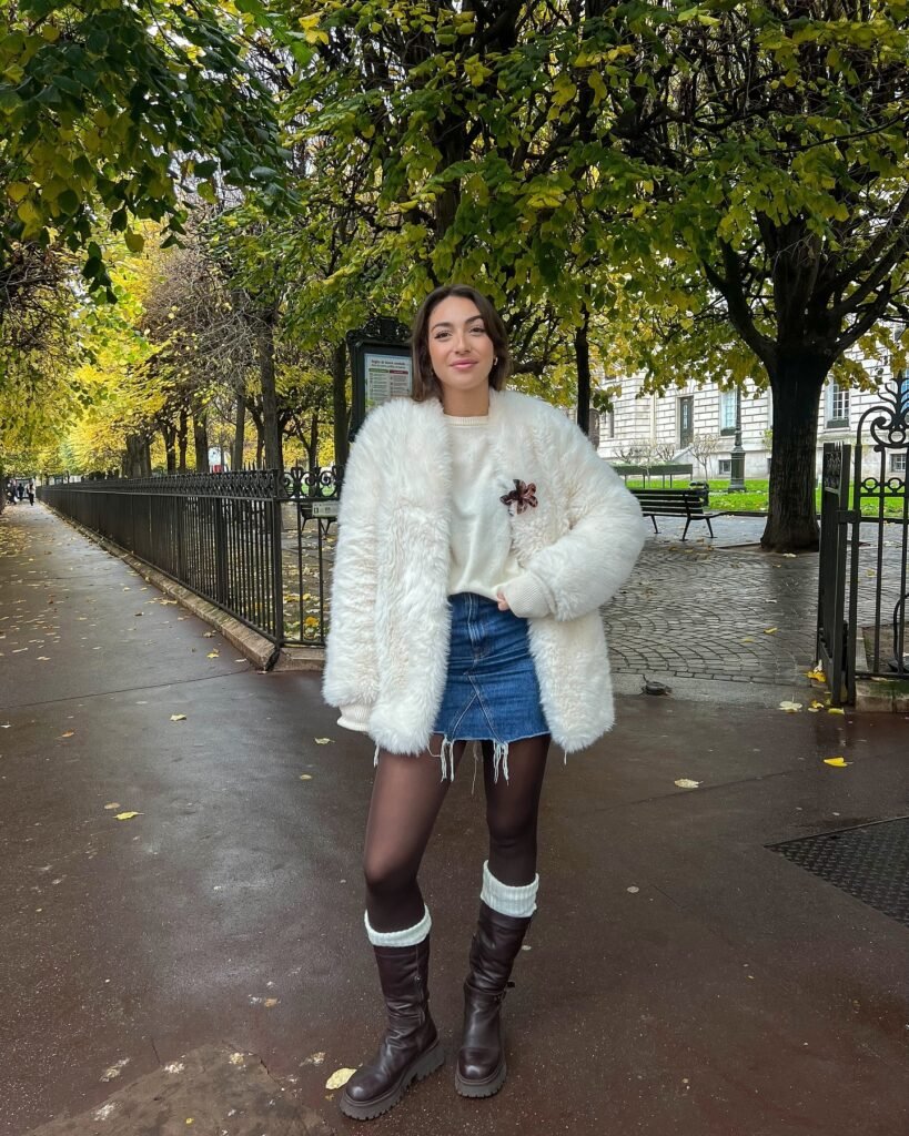 A stylish woman wearing a denim mini skirt, white fluffy coat, tights, and knee-high boots, standing in a park during winter, showcasing a trendy cold-weather outfit.