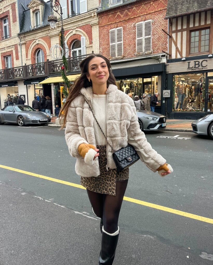 A woman walking on a city street wearing a leopard-print mini skirt, faux fur coat, black tights, knee-high boots, and carrying a quilted crossbody bag.